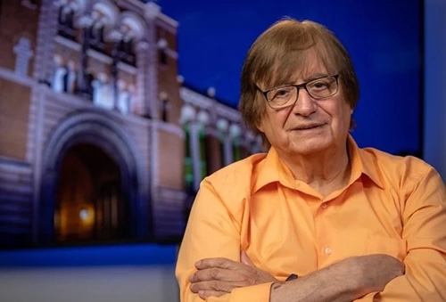 Richard Tapia wearing an orange shirt with his arms crossed, sitting in front of an academic building.