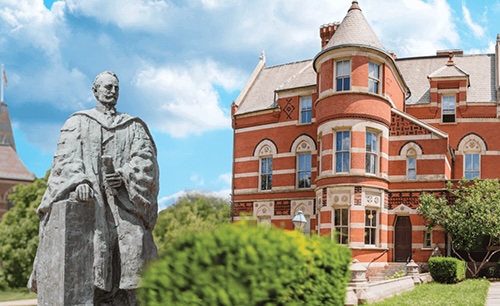 A statue of a man in doctorate robs in the foreground, and a red brick castle-like building in the background.