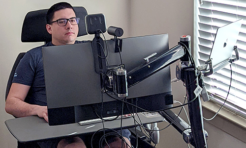 A computing student sits at his desk where he has multiple screens and technology set up.