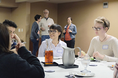 Young people sit around a table talking. There is food and drinks on the table. Educators and mentors are seen talking in the background.