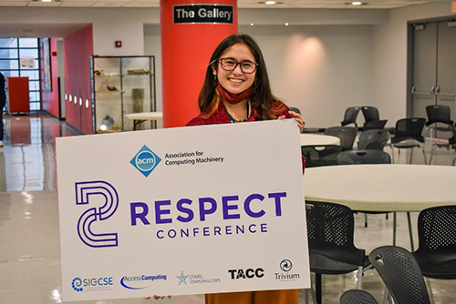Cole is photographed holding up a RESPECT conference sign. They are a filipina, female-presenting person with long brown hair and pink glasses, and they are grinning at the camera. The sign she is holding is so big it covers the rest of her body in frame.