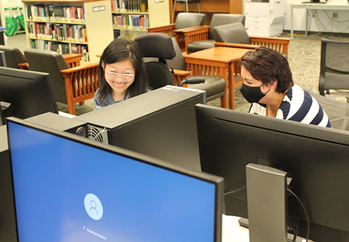 Two students in a library on campus smiling while using computers