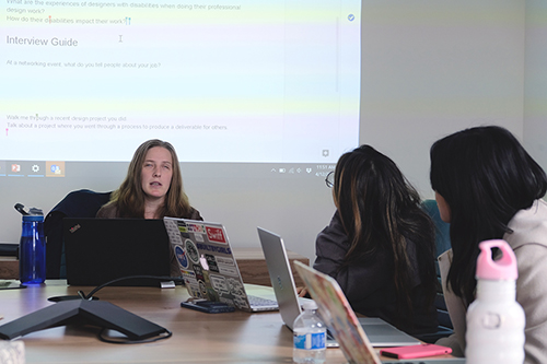 A blind instructor sits in front of a powerpoint talking to two students, all with laptops open.