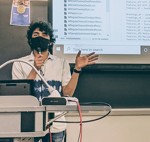 A graduate student in a mask speaks into a microphone; they stand in front of a classroom podium and a projector featuring files and code.