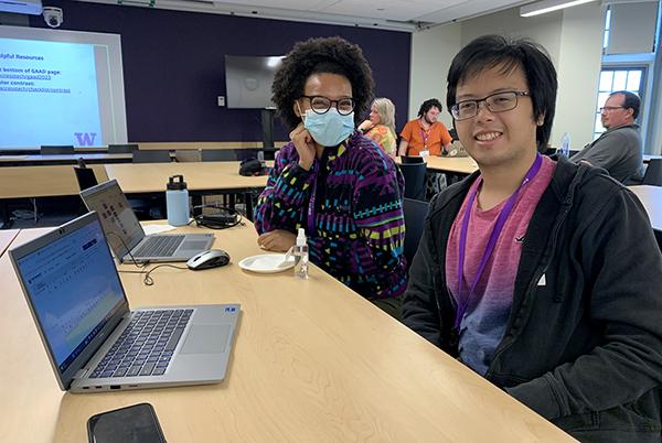 Two educators smile for the camera while working on a laptop together.