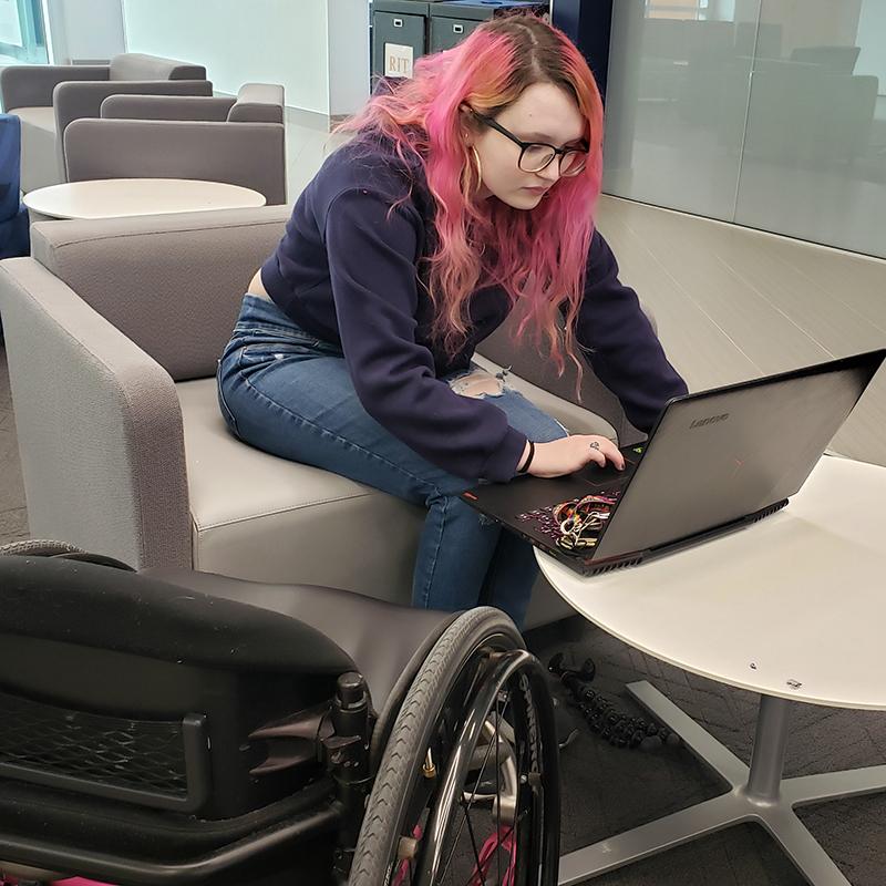 An AccessComputing student sits in a chair and types on a computer, her wheelchair off to the side.