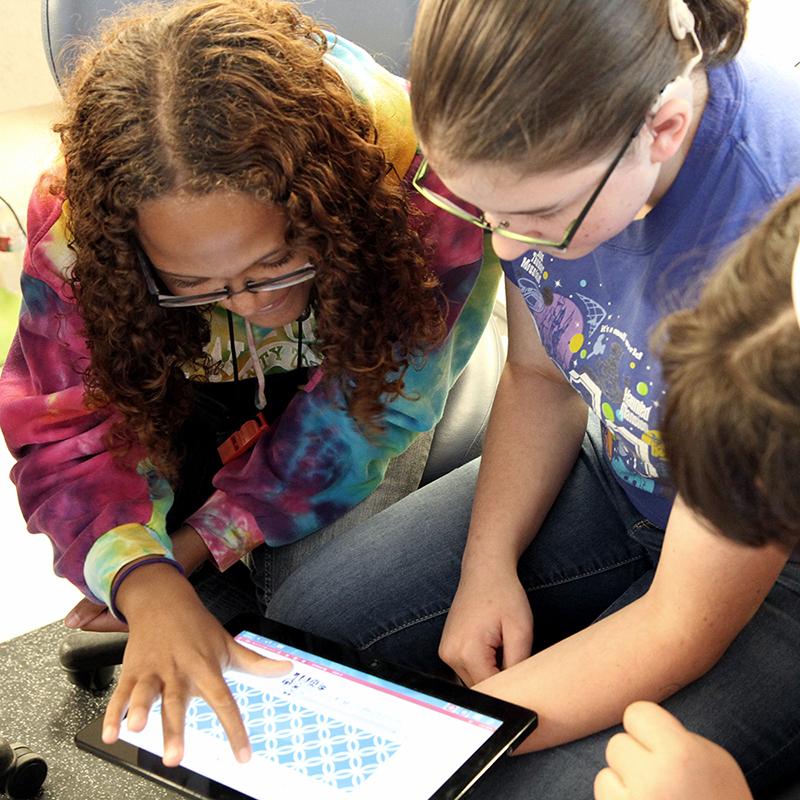 Three students look at a tablet together; one student enlarging the screen with her fingers.