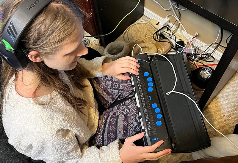A blind student uses a Braille display and headphones while sitting at her desk.